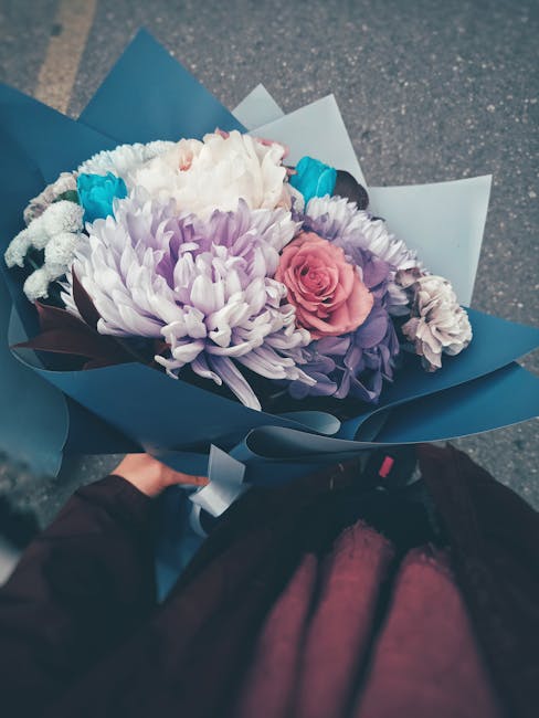 A hand holding a beautifully arranged bouquet of fresh flowers wrapped in elegant blue and white paper. The bouquet features a mix of flowers including a large white chrysanthemum, pink roses, and pur