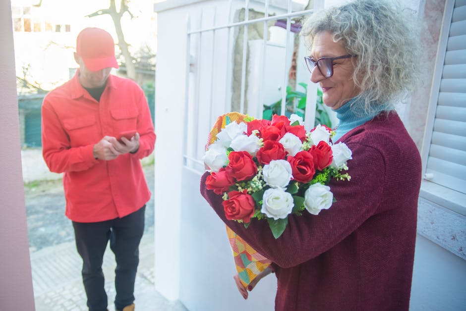 An elderly woman with curly gray hair, wearing glasses, a light blue turtleneck, and a maroon cardigan, is holding a floral bouquet composed of red and white roses arranged with green foliage in a col