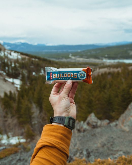 A person's hand, wearing a black wristwatch and a mustard-colored jacket sleeve, holds a blue and orange 'Builders Protein' energy bar against a scenic outdoor landscape. The background features rolli